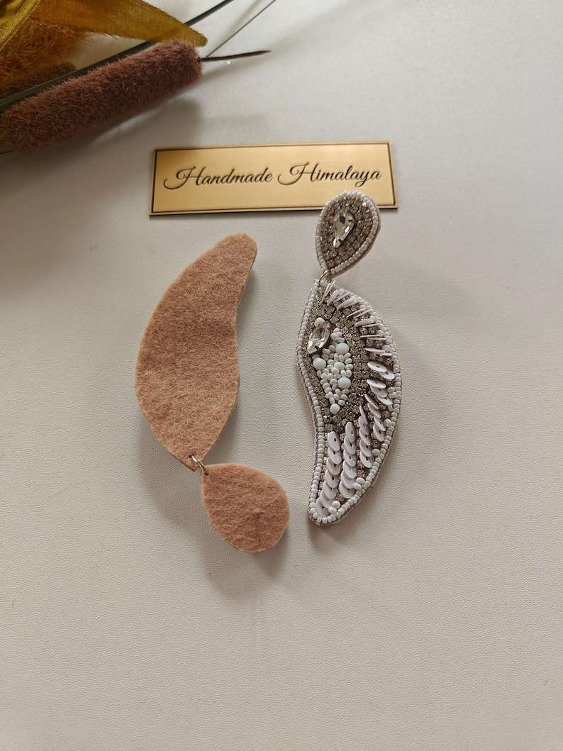 Op de afbeelding: Een paar oorbellen met een halvemaanvormig ontwerp. De ene oorbel toont een achterkant van beige vilt, terwijl de andere ingewikkeld kralenwerk met witte pailletten, heldere kristallen en zilverkleurige kralen bevat. Een gouden label vermeldt Handmade Himalaya.