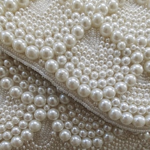 Extreme macro close-up focusing on the exquisite texture of the ivory pearls and seed beads under soft, diffused light. The varying diameters of the pearls create a three-dimensional landscape of shimmer and shadow. This shot highlights the meticulous hand-stitching and the high-grade iridescent finish of the materials, serving as a testament to the artisan quality of the piece.