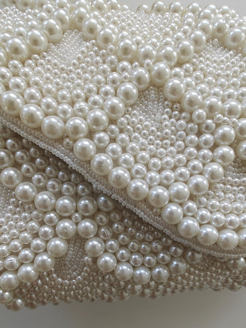 Extreme macro close-up focusing on the exquisite texture of the ivory pearls and seed beads under soft, diffused light. The varying diameters of the pearls create a three-dimensional landscape of shimmer and shadow. This shot highlights the meticulous hand-stitching and the high-grade iridescent finish of the materials, serving as a testament to the artisan quality of the piece.