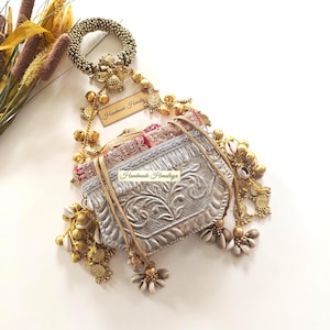 以下が含まれることがあります： 複雑な刺繍、メタリックなフリンジ、ビーズのハンドルが特徴のシルバーカラーの巾着袋。バッグには金色の鈴、小さなコイン、白い貝殻があしらわれています。長方形のタグには「Handmade Himalaya」という文字が表示されています。