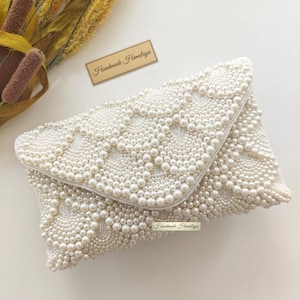 A top-down flat lay view showcases the bridal handbag resting on a clean white surface alongside textural dried botanicals. The scalloped envelope flap is the focal point, revealing the precision of the beaded borders and the undulating wave motifs. This angle highlights the rich, creamy ivory color palette and the sophisticated symmetry that defines this luxury wedding accessory.