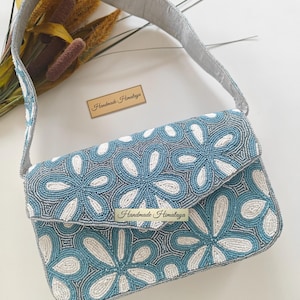 This high-angle flat lay features the Blue Beaded Bag alongside decorative botanicals and a brand card to emphasize its artisan beaded purse heritage. The Portuguese Azulejos inspired patterns are displayed clearly across the rectangular frame. In terms of scale, the bridal shower accessory is laid next to natural pampas grass, showing it is a mid-sized evening shoulder bag perfectly proportioned to serve as a standout wedding guest bag.