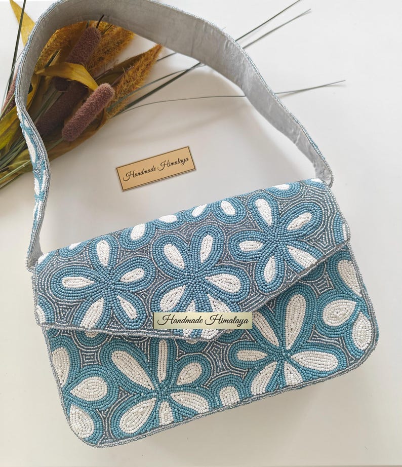 This high-angle flat lay features the Blue Beaded Bag alongside decorative botanicals and a brand card to emphasize its artisan beaded purse heritage. The Portuguese Azulejos inspired patterns are displayed clearly across the rectangular frame. In terms of scale, the bridal shower accessory is laid next to natural pampas grass, showing it is a mid-sized evening shoulder bag perfectly proportioned to serve as a standout wedding guest bag.