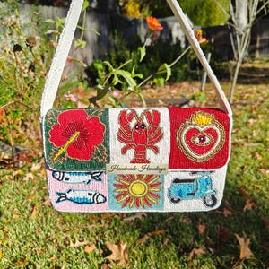 May include: A white beaded shoulder bag with a rectangular flap, featuring colorful beaded designs. The designs include a red hibiscus flower, a lobster, a heart with an eye, fish, a sun, and a scooter. The text "Handmade Himalaya" is visible.