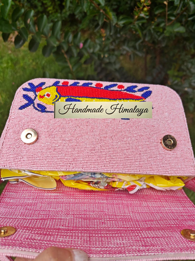 Interior view of the open pink quirky handbag revealing the magnetic closure and pink fabric lining. This handmade Indian beaded bag shows the single compartment currently filled with packing material to maintain its shape. The artisan beaded bag construction combines canvas and jute with high-quality hardware. This view emphasizes the functionality of the small clutch bag, designed to keep essentials safe while maintaining its coastal aesthetic.