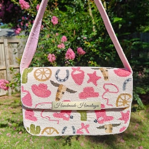 May include: A beaded crossbody bag with a pink strap and a flap closure. The bag is off-white with pink, gold, and green cowboy-themed designs. The text "Handmade Himalaya" is on a metal plate on the front.