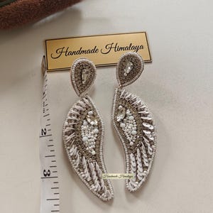 Op de afbeelding: Een paar wit-zilveren kralenoorbellen met druppelvormige bovenkanten en gebogen, bladachtige hangers. De oorbellen zijn voorzien van heldere kristallen, witte kralen en lovertjes. Een meetlint toont een lengte van ongeveer 8,9 cm.