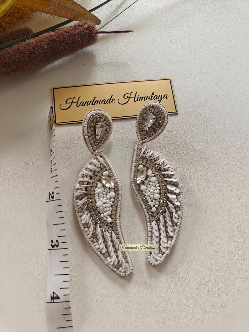 Op de afbeelding: Een paar wit-zilveren kralenoorbellen met druppelvormige bovenkanten en gebogen, bladachtige hangers. De oorbellen zijn voorzien van heldere kristallen, witte kralen en lovertjes. Een meetlint toont een lengte van ongeveer 8,9 cm.