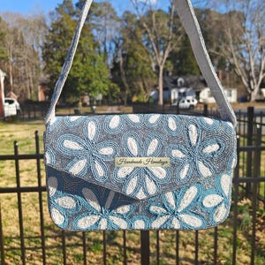 This front view showcases the Dusty Blue Floral Beaded Shoulder Bag suspended against a natural garden backdrop to highlight its organic Chinoiserie style. The intricate floral beadwork features ivory and cornflower blue blooms hand-sewn onto a shimmering silver-grey base. For scale, this Something Blue Bridal Clutch is approximately the width of a standard tablet (approx 7 inches), offering a sophisticated silhouette that remains lightweight and portable.