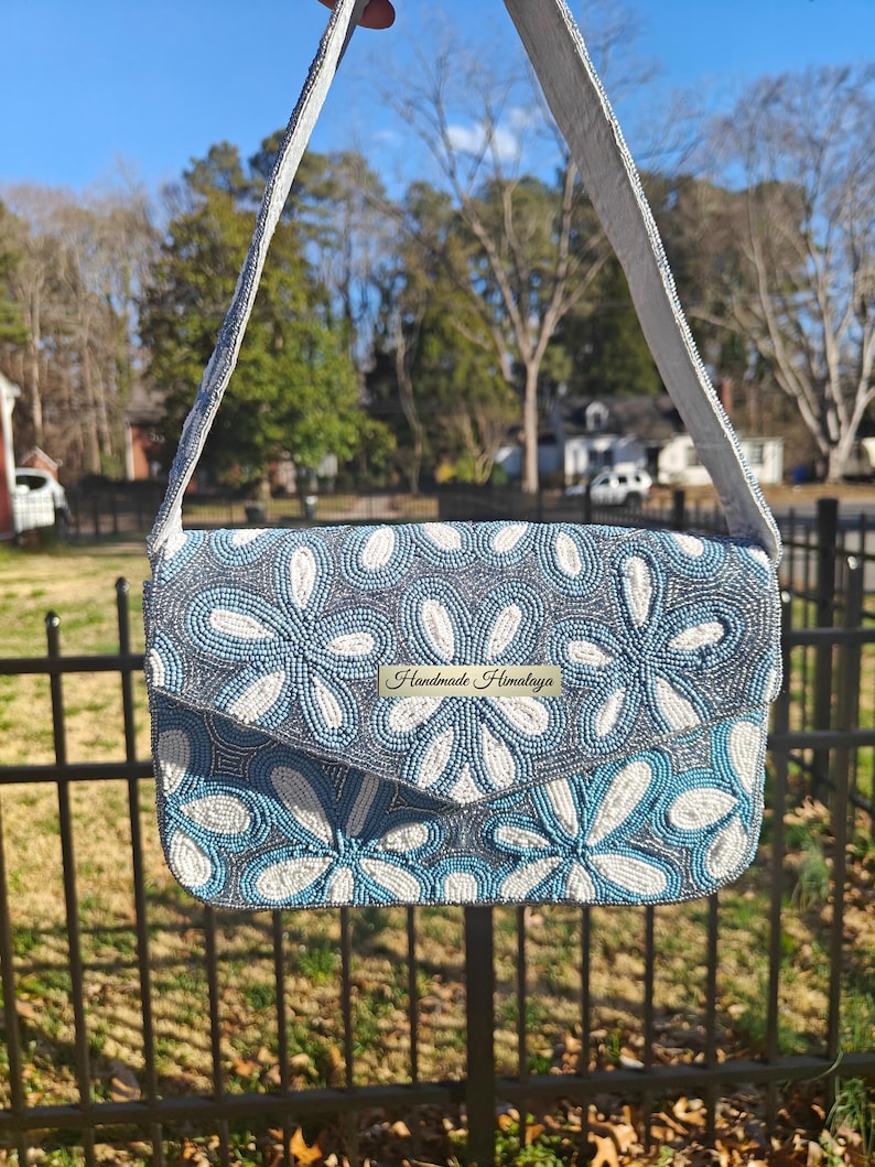 This front view showcases the Dusty Blue Floral Beaded Shoulder Bag suspended against a natural garden backdrop to highlight its organic Chinoiserie style. The intricate floral beadwork features ivory and cornflower blue blooms hand-sewn onto a shimmering silver-grey base. For scale, this Something Blue Bridal Clutch is approximately the width of a standard tablet (approx 7 inches), offering a sophisticated silhouette that remains lightweight and portable.