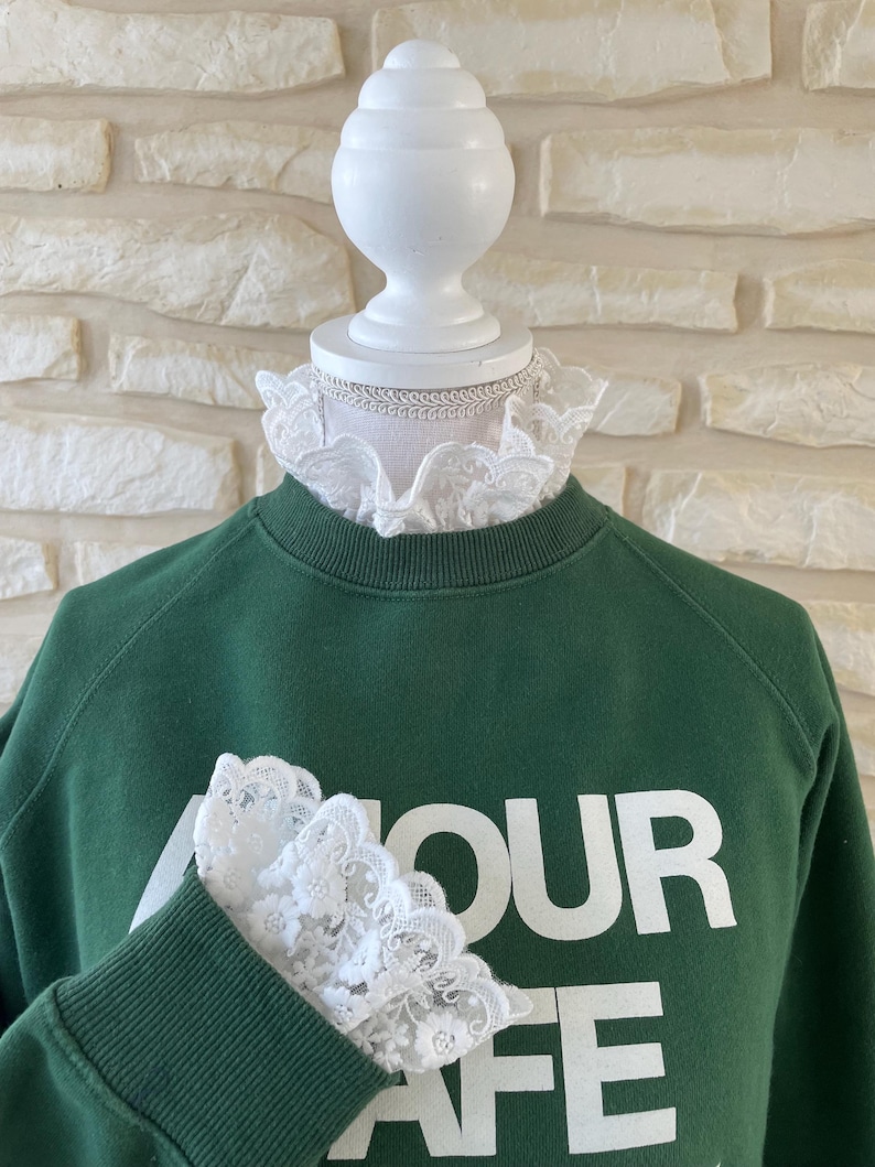 May include: A green sweatshirt with the text "Amour Cafe" printed on the front. The sweatshirt has white lace trim around the neck and on the sleeves.