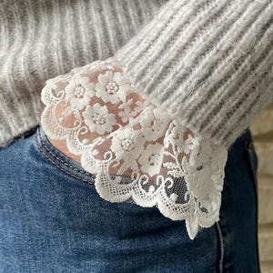 May include: A gray knitted sweater with a white lace trim on the cuffs. The sweater is worn over a pair of blue jeans.