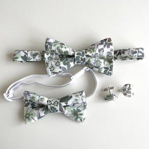 May include: A set of matching bow ties and cufflinks. The bow ties feature a floral pattern in shades of green, white, and gray. The set includes an adult bow tie, a child's bow tie, and a pair of cufflinks with the same floral design.