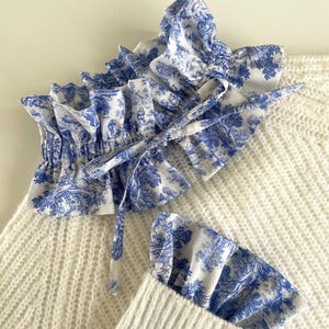 Könnte beinhalten: Ein weißer Strickpullover mit einem blau-weißen Toile-de-Jouy-Print-Kragen und -Manschetten. Der Kragen ist gerüscht und mit einer passenden Stoffschleife gebunden. Die Manschetten sind ebenfalls gerüscht.