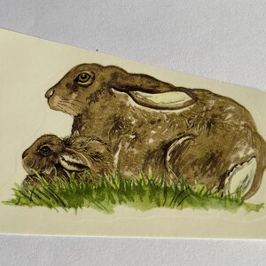 Op de afbeelding: Een aquarel van een bruine haas en een babyhaas die in een stukje groen gras zitten.