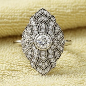 May include: A silver ring with an Art Deco design. The ring features a large, central, round diamond surrounded by smaller diamonds in an ornate, symmetrical pattern. The ring is set against a yellow background.