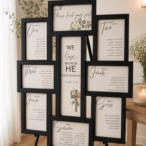 May include: A black multi-frame collage display on an easel featuring white seating chart cards for tables one through seven. The center panel displays the text "WE love BECAUSE HE FIRST LOVED US 1 John 4:19" with a floral cross illustration.