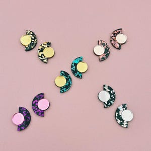 Puede incluir: Una colección de diez pendientes semicirculares con base negra y patrones moteados multicolores. Cada pendiente incluye un acento metálico circular en oro, plata, oro rosa o rosa, unido al borde plano de la forma de medio círculo.