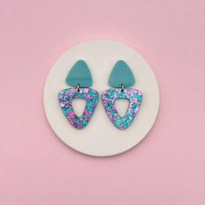 May include: A pair of teal and iridescent pink triangle earrings. Each earring has a teal triangle stud and a larger open triangle with a glittery design. The earrings are displayed on a white circular surface against a pink background.