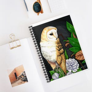 May include: Spiral-bound notebook featuring a detailed illustration of a barn owl with white, tan, and brown feathers. The owl is perched among green leaves and white flowers against a black background. The notebook is open on a white surface.