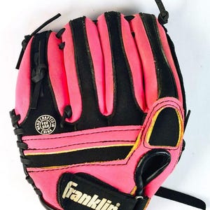 May include: A pink and black baseball glove with the brand name "Franklin" visible. The glove has black stitching and accents, with a yellow trim. The words "Hand Crafted" and "China" are printed on a circular emblem.