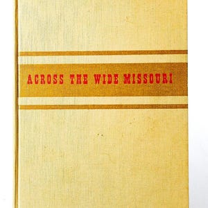 May include: A hardcover book with a light yellow, textured fabric cover. A horizontal gold band spans the center, featuring the title ACROSS THE WIDE MISSOURI printed in bold red capital letters. The book has a rectangular shape and a vertical spine.
