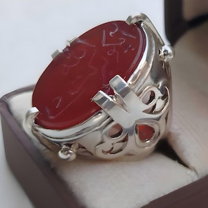May include: A silver ring featuring a large, oval-shaped, deep red stone with intricate engraved calligraphy on its surface. The metal band displays an openwork, decorative pattern with polished silver prongs securing the central gemstone.