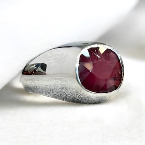 Peut inclure: Une bague en argent avec une grande pierre précieuse rouge foncé. La bague a un dessus arrondi et plat et une finition lisse et polie.