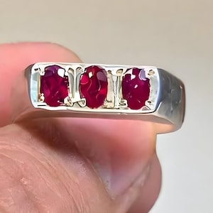 Peut inclure: Une bague en argent ornée de trois pierres précieuses ovales rouge foncé. Les pierres sont serties dans une lunette dorée, créant un contraste saisissant avec le corps en argent. La bague est tenue par une main, mettant en valeur son design.