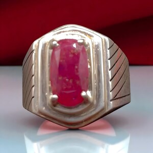 Könnte beinhalten: Ein silberner Ring mit einem großen, oval geschliffenen Rubin. Der Rubin hat eine leuchtend rote Farbe mit inneren Einschlüssen. Der Ring hat ein geometrisches Design mit geätzten Details an den Seiten, was einen klassischen Stil zeigt.