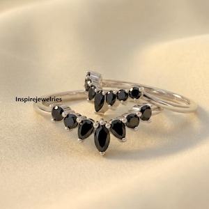Può includere: Due anelli d'argento con pietre nere disposte a forma curva. Le pietre sono rotonde e a goccia. Gli anelli sono su uno sfondo bianco sporco. Il testo "Inspirejewelries" è visibile.