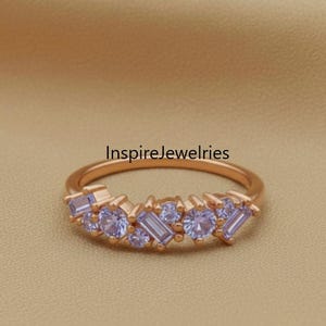 以下が含まれることがあります： 様々な形のライトブルーの宝石がクラスターになった、ローズゴールドのリング。リングは繊細なバンドで、「Inspirejewelries」の文字が見える。