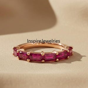 Peut inclure: Une bague en or rose ornée de pierres précieuses rectangulaires rose foncé disposées autour du cercle. La bague est présentée sur une surface aux tons neutres. Le texte "InspireJewelries" est visible.