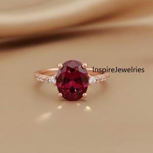 Może przedstawiać: Pierścionek z różowego złota z dużym, owalnym, ciemnoczerwonym kamieniem szlachetnym. Oprawa ozdobiona jest mniejszymi, przezroczystymi kamieniami. Pierścionek jest prezentowany na neutralnym tle, podkreślając jego elegancki design.