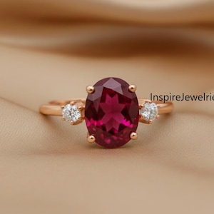 Op de afbeelding: Rosegouden ring met een grote, ovaal geslepen, dieprode edelsteen als middelpunt, geflankeerd door twee kleinere, ronde, heldere edelstenen. De ring is geplaatst tegen een neutrale, bruinachtige achtergrond. De tekst "InspireJewelries" is zichtbaar.