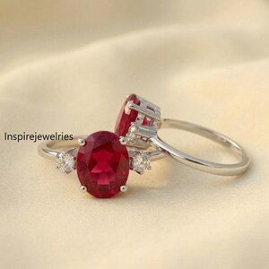 May include: Two silver rings featuring large oval, deep red gemstones. Each ring has smaller, round, clear gemstones flanking the main stone. The rings are set against a soft, cream-coloured background. The text "Inspirejewelries" is visible.