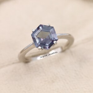 May include: A silver ring featuring a hexagonal, iridescent purple gemstone. The ring is set in a simple band and has a delicate design. "Inspire Jewelries" is printed on the band.