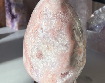Puede incluir: Una gran geoda de ágata rosa y blanca con un exterior natural y rugoso. La geoda está sobre un soporte de madera.