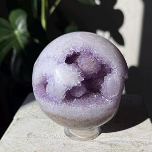 May include: A spherical amethyst geode, showcasing a light purple exterior and a cavity filled with sparkling crystal formations. The geode sits on a clear stand, with a neutral background.