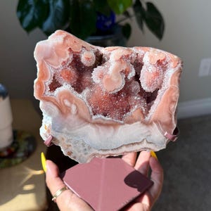 May include: A large, pink and white geode crystal with a sparkling interior. The geode is held above a pink, square-shaped stone. The crystal is displayed on a metal stand. The geode is a natural, decorative item.