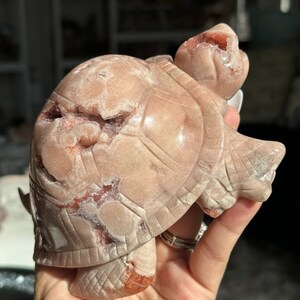 May include: A carved pink and red agate turtle figurine. The turtle is standing on its four legs with its head raised. The agate has a natural crystal formation with a sparkly finish.