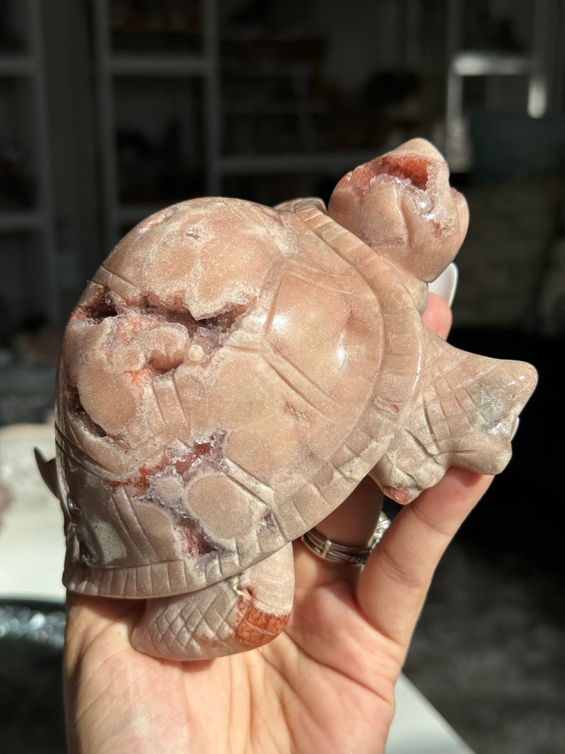 May include: A carved pink and red agate turtle figurine. The turtle is standing on its four legs with its head raised. The agate has a natural crystal formation with a sparkly finish.