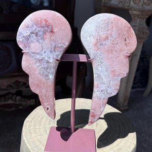 May include: A pair of pink and white crystal angel wings mounted on a mauve metal stand. The wings have a rough, textured surface and are displayed on a light-colored, round pedestal. The wings are a decorative item.