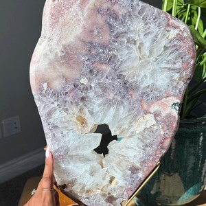 May include: A large, irregularly shaped pink and white crystal geode slice. The geode has a hollow center and is mounted on a gold-colored metal stand. The crystal's interior features a crystalline structure.