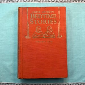 May include: A bright orange hardcover book titled Uncle Arthur's Bedtime Stories, Volumes 15-16. The front cover features gold embossed text and a line drawing of two children reading, flanked by two tall, thin candle illustrations on each side.