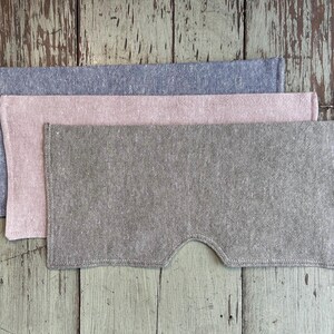 May include: Three linen eye masks in shades of gray, pink, and green are stacked on a weathered wooden surface. The bottom mask has a curved cutout for the nose. The masks are made of a textured fabric.