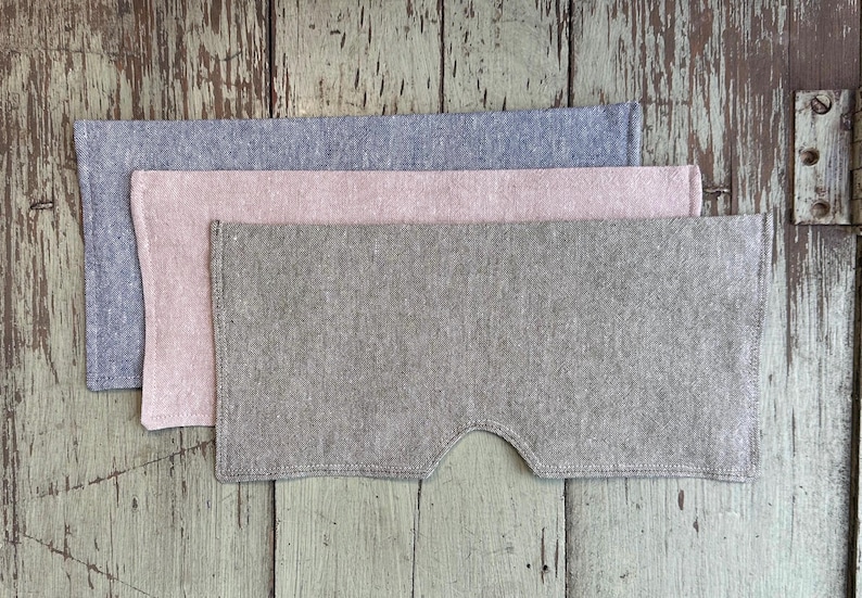 May include: Three linen eye masks in shades of gray, pink, and green are stacked on a weathered wooden surface. The bottom mask has a curved cutout for the nose. The masks are made of a textured fabric.