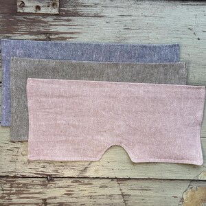 May include: Three linen eye masks in shades of blue, gray, and pink are stacked on a weathered wooden surface. The pink mask has a curved cutout for the nose. The fabric has a textured appearance.