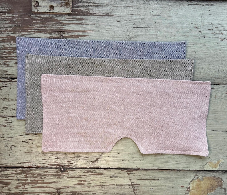 May include: Three linen eye masks in shades of blue, gray, and pink are stacked on a weathered wooden surface. The pink mask has a curved cutout for the nose. The fabric has a textured appearance.