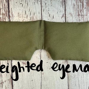 May include: An olive green weighted eye mask. The mask is rectangular with a curved cutout for the nose. The words "weighted eye mask" are written in black script below the mask. The mask is made of a soft fabric.